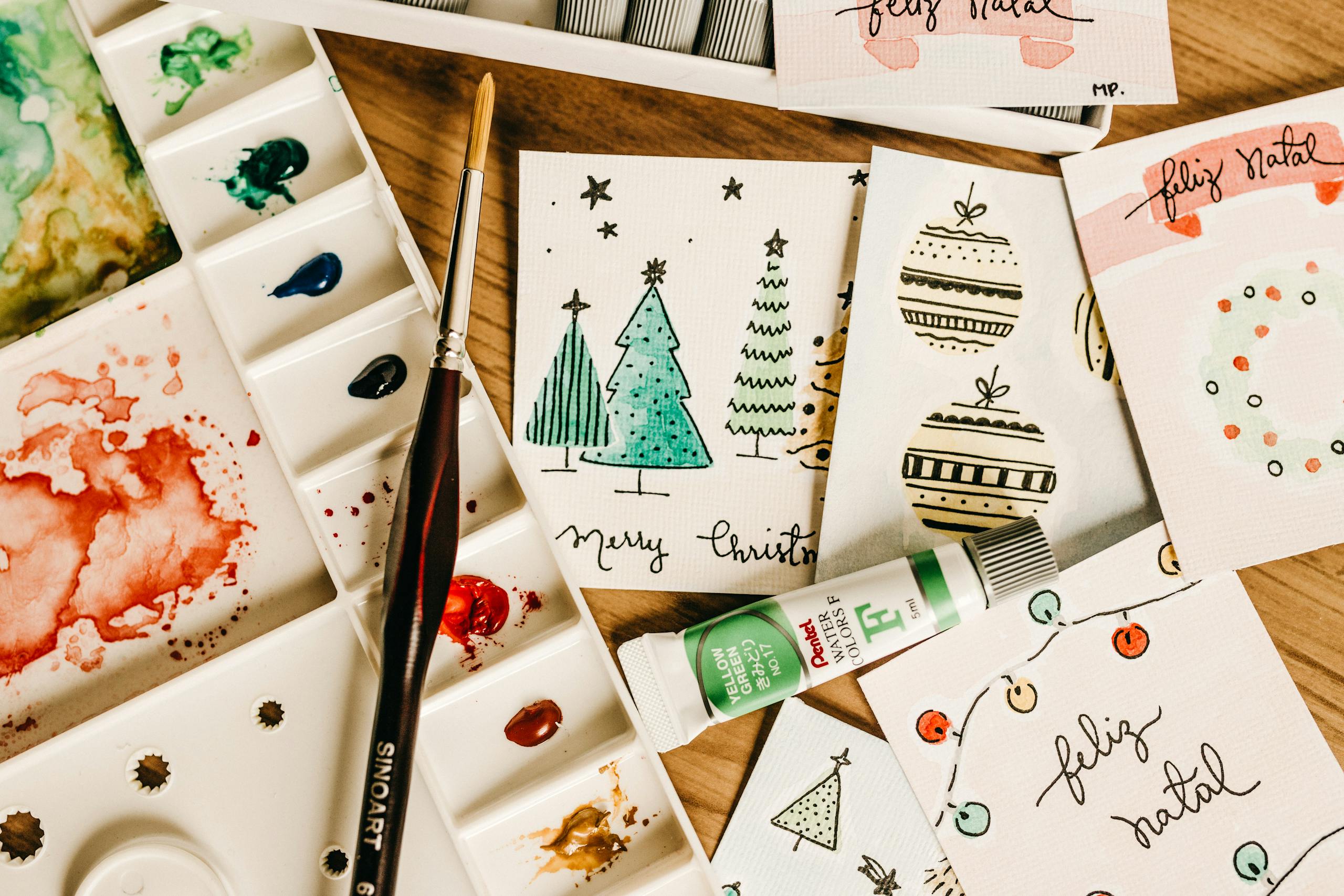 Artistic holiday card designs with watercolor paints and brushes on a wooden table.