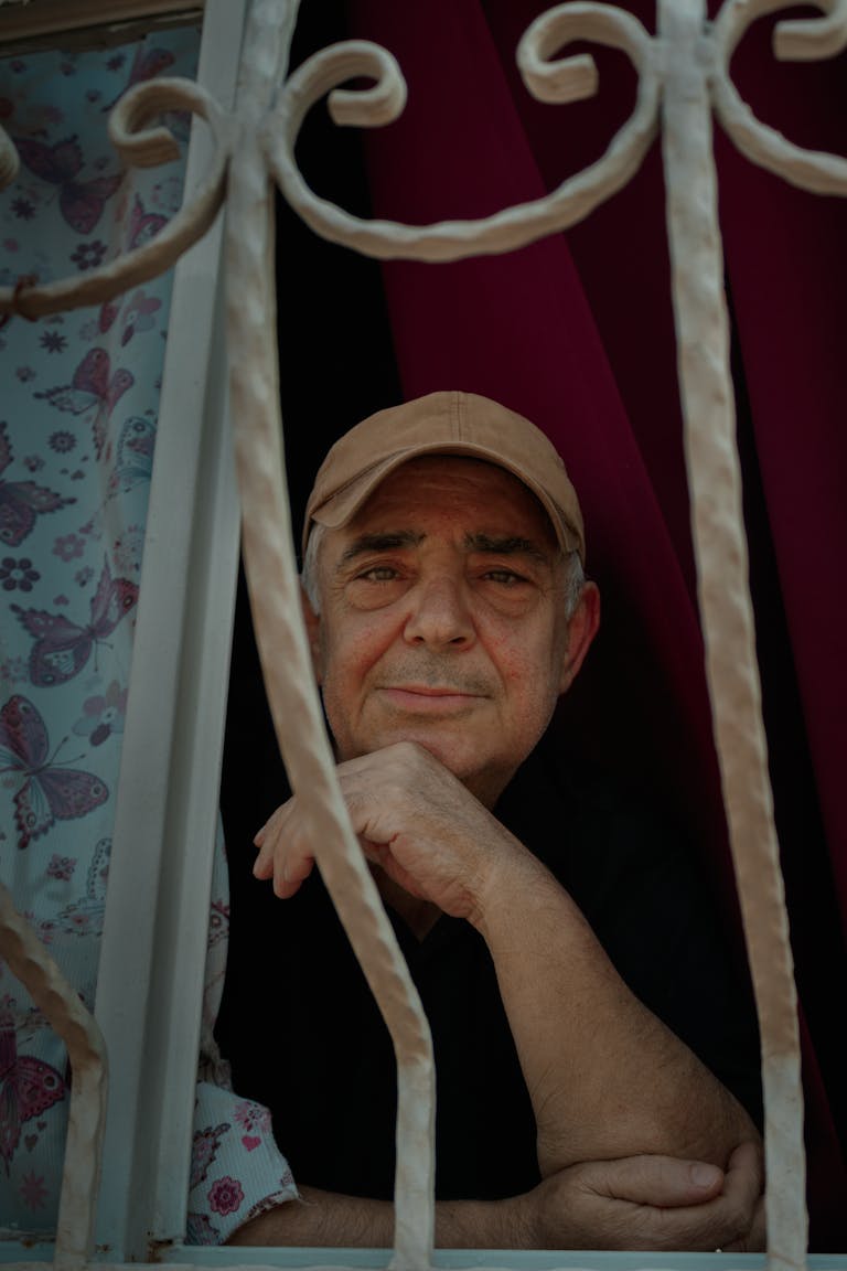 Elderly man with cap gazing from window with floral curtain, exuding calmness.