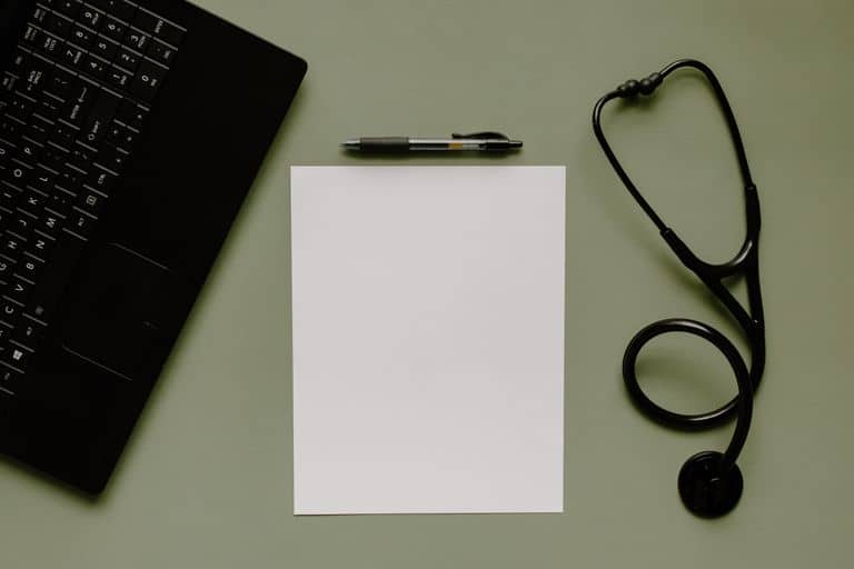 Medical workspace featuring a blank paper, stethoscope, and laptop from above.
