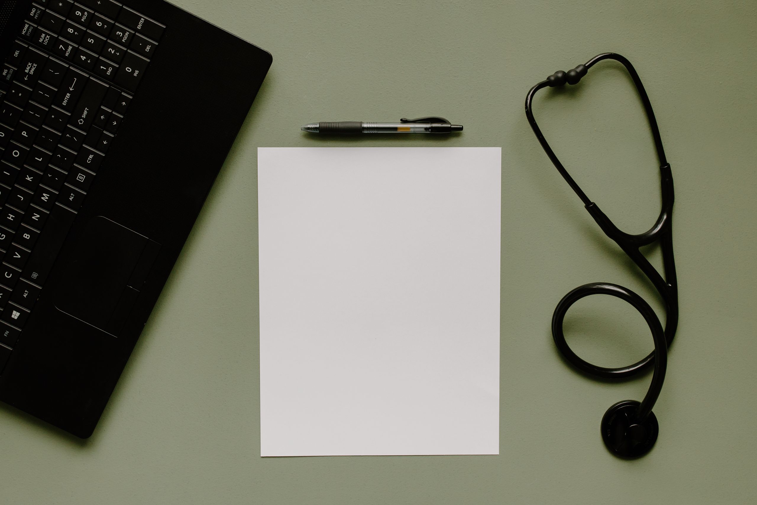 Medical workspace featuring a blank paper, stethoscope, and laptop from above.